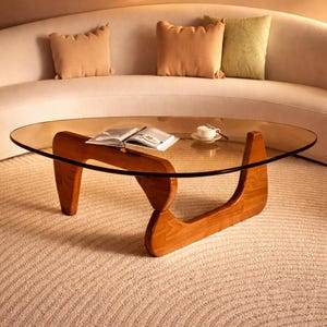 May include: A mid-century modern coffee table with a clear oval glass top and a wooden base. The base is made of two curved wooden supports. A book and a cup of coffee sit on the table. The table is in a living room setting.