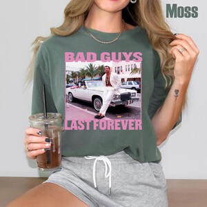 May include: Moss green t-shirt with a graphic featuring a man in a white suit leaning on a white convertible car. The text "BAD GUYS" and "LAST FOREVER" are in pink letters.