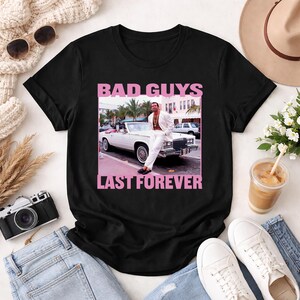 May include: Black t-shirt with a pink graphic that reads "BAD GUYS LAST FOREVER." The graphic features a man in a white suit leaning against a white convertible car. The image has a retro aesthetic.