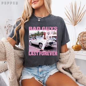 May include: Dark gray t-shirt with a pink graphic that reads "BAD GUYS LAST FOREVER." The graphic features a man in a white suit leaning against a white convertible. The shirt has a vintage, worn look.