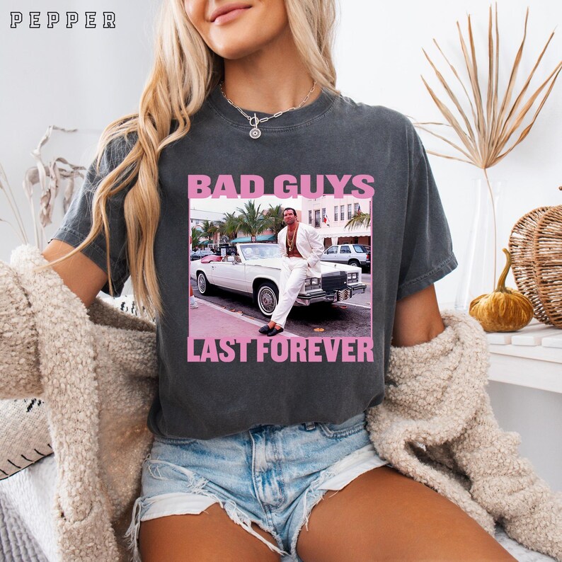 May include: Dark gray t-shirt with a pink graphic that reads "BAD GUYS LAST FOREVER." The graphic features a man in a white suit leaning against a white convertible. The shirt has a vintage, worn look.