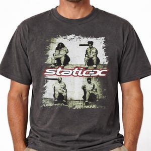 May include: Dark gray t-shirt featuring a graphic with four seated figures and the band name "Static-X" in red and white. The design has a distressed, vintage look. The shirt has short sleeves and a crew neck.