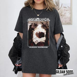 May include: Dark heather gray t-shirt featuring a graphic with the word "Dystopia" above an image of a primate holding a heart-shaped object. The text "HUMAN=GARBAGE" is below the image. The shirt is a Gildan 5000 Dark Heather.