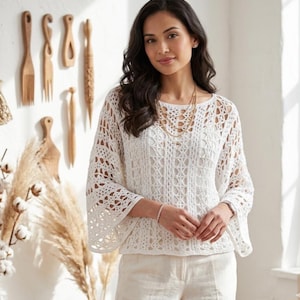 May include: A white crochet top featuring an intricate openwork pattern, a wide boat neckline, and loose, flared three-quarter length sleeves. The garment is constructed with a repeating geometric lace design and is worn over a solid white base layer.