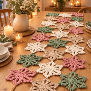 Puede incluir: Un camino de mesa compuesto por motivos de copos de nieve tejidos a ganchillo e interconectados en tonos verde salvia, rosa empolvado, crema y beige. El camino se extiende sobre una mesa de madera con un patrón floral repetitivo y calado.