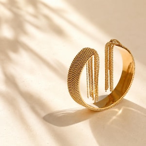 Peut inclure: Un bracelet manchette doré composé d'une large bande à la surface perlée et texturée. Une extrémité de la manchette est ornée de multiples chaînes dorées fines et pendantes qui tombent du bord, créant un aspect fluide et superposé.