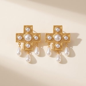 Può includere: Un paio di orecchini a forma di croce color oro con perle finte. Ogni orecchino presenta una grande perla centrale, circondata da perle più piccole e tre perle a goccia pendenti. Gli orecchini hanno un bordo strutturato.