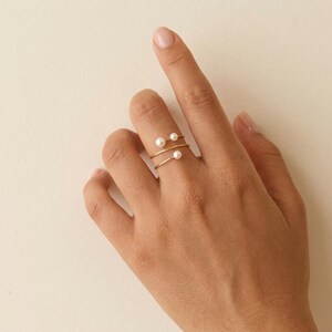 May include: A gold-coloured ring with a spiral design, featuring two small, white pearls. The ring is worn on a finger, showcasing its delicate and minimalist style. The ring's design is simple and elegant.