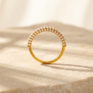 Puede incluir: Un anillo dorado con dos filas de pequeñas perlas blancas. El anillo tiene una banda de oro pulido y las perlas están espaciadas uniformemente en la mitad superior. El anillo se muestra sobre una superficie clara.