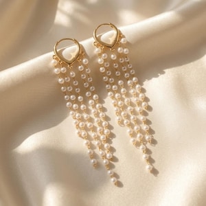 May include: Gold-tone hoop earrings with a teardrop detail, featuring multiple strands of small, white pearls. The earrings are presented on a cream-coloured, satin fabric, creating a luxurious appearance.