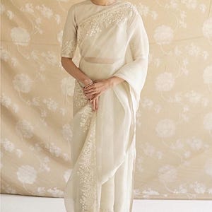 May include: A cream-colored saree featuring intricate white floral embroidery along the border and shoulder. The garment includes a short-sleeved blouse and a sheer, draped fabric that falls to the floor, showcasing detailed needlework throughout.