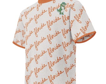 May include: A light gray short-sleeve t-shirt featuring an all-over repeating pattern of the word florida written in orange cursive script. The shirt has an orange ribbed crew neckline and orange sleeve cuffs, with a small green alligator graphic.