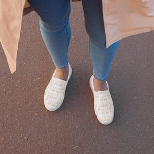 May include: White slip-on shoes with a repeating tan pattern. The shoes are worn with blue jeans and a tan coat. The shoes appear to be made of canvas or a similar material.