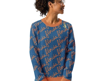 May include: A blue long-sleeve sweatshirt featuring a repeating diagonal pattern of the word florida written in orange cursive script, with a small green alligator graphic on the upper chest and orange ribbed trim at the neck, cuffs, and hem.