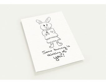 May include: A white greeting card featuring a black line drawing of a rabbit wearing a sweater and skirt while holding a carrot. Below the illustration, the handwritten text reads: Some bunny is thinking of you!