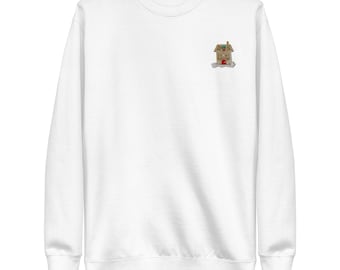 May include: A white long-sleeve crewneck sweatshirt featuring a small embroidered design on the upper left chest. The embroidery depicts a brown gingerbread house with a red door, a green wreath above the door, and white icing details on snow.