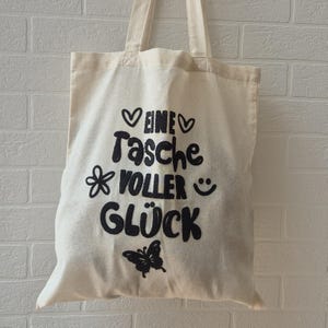 Könnte beinhalten: Eine cremefarbene Stofftasche mit zwei Tragegriffen, die mit dem schwarzen Schriftzug EINE TASCHE VOLLER GLÜCK bedruckt ist, ergänzt durch schwarze grafische Symbole von zwei Herzen, einer Blume, einem Smiley und einem Schmetterling.