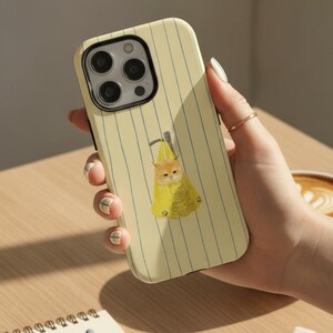 May include: A pale yellow smartphone case with thin vertical blue stripes. The centre of the case features a graphic illustration of a small orange kitten sitting inside a yellow plastic shopping bag with a handle at the top.