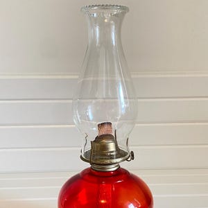 Op de afbeelding: Een vintage olielamp met een heldere glazen schoorsteen en een levendige rode glazen voet. De lamp heeft een messing brander en een klassiek ontwerp, perfect om een vleugje rustieke charme toe te voegen.
