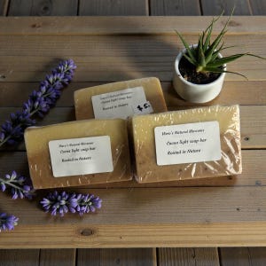Puede incluir: Tres jabones de cacao de Viero's Natural Blossoms, envueltos en plástico transparente. Cada barra tiene un color de dos tonos, con una parte superior clara y una inferior más oscura. Las barras están etiquetadas como "Cocoa light soap bar" y "Rooted in Nature". También hay una pequeña planta en maceta y ramitas de lavanda.