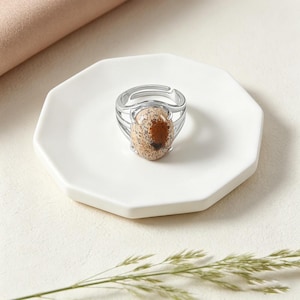 May include: A silver-toned ring with an oval, speckled stone. The stone features a mix of beige, brown, and dark brown hues. The ring's band has a multi-layered design. The ring is displayed on a white, octagonal plate.