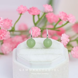 Puede incluir: Un par de pendientes redondos de piedra preciosa verde claro con ganchos plateados. Los pendientes se exhiben en una caja de joyería blanca, con flores rosas en el fondo. El texto "GEMORASTUDIO" y "Handcrafted Jewellery" son visibles.