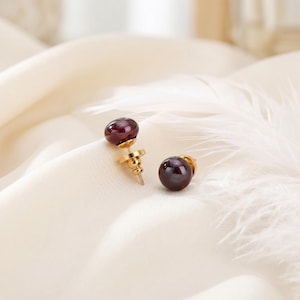 May include: A pair of stud earrings featuring a dark red, spherical gemstone set on gold posts. The earrings are displayed on a soft, white fabric with a feather in the background. The earrings have a classic, elegant design.