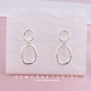 Może przedstawiać: Para srebrnych kolczyków o podwójnym wzorze pętli. Kolczyki mają małą, okrągłą górną pętlę połączoną z większą, owalną dolną pętlą. Sekcja łącząca jest ozdobiona małymi, błyszczącymi kamieniami. Tekst "GEMORAS STUDIOL Handcrafted Jewellery" znajduje się na dole.