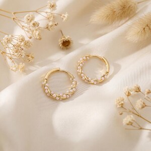 May include: A pair of gold-colored hoop earrings, each adorned with small, white pearls. The earrings have a twisted design and are set against a cream-colored fabric, with dried flowers in the background.