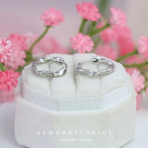 Puede incluir: Un par de pendientes de aro plateados con un diseño retorcido, cada uno adornado con pequeñas piedras brillantes. Los pendientes descansan sobre una superficie de terciopelo blanco, con flores rosas en el fondo. El texto "GEMORASTUDIOL Handcrafted Jewellery" es visible.