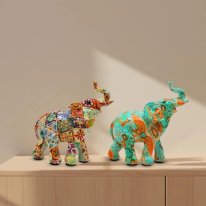 May include: Two decorative elephant figurines. One is a patchwork of colorful floral and geometric patterns. The other is turquoise with orange floral designs. Both have raised trunks and are displayed on a wooden surface.