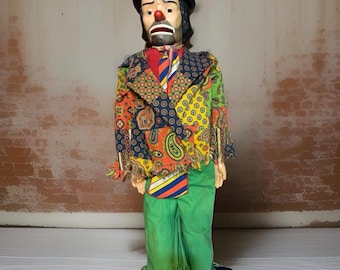 May include: A ventriloquist dummy clown figure wearing a patchwork jacket with paisley and geometric patterns in orange, yellow, green, and blue. The clown has a red nose, black hair, a black hat, a striped necktie, and solid green trousers with fringe.