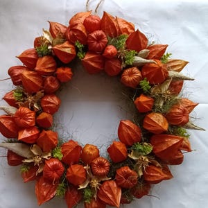 Könnte beinhalten: Ein kreisförmiger Kranz aus zahlreichen leuchtend orangefarbenen Physalis-Hüllen, auch Lampionblumen genannt. Das Arrangement ist mit kleinen Büscheln aus grünem Blattwerk und getrockneten, blassen, spitzen Samenkapseln auf einer Basis befestigt.