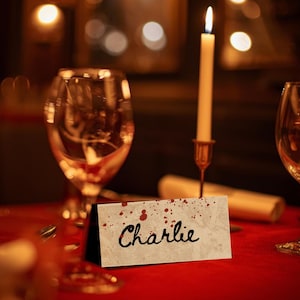 May include: A dimly lit table setting with a red tablecloth, two wine glasses, and a lit candle in a brass holder. A name card with the name "Charlie" is displayed, with red splatter details.