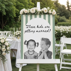 Op de afbeelding: Een wit houten bord met de tekst "These kids are getting married!" toont een foto van twee lachende kinderen. Het bord is versierd met witte en groene strepen en bekroond met witte rozen. Het bord staat op een bruiloft in de buitenlucht.