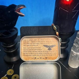 May include: An open, silver-colored metal tin with a printed message about crows, a black raven statue, a black candle, and three gold coins. The text on the tin reads, "Crows are intelligent, playful, and endlessly curious."