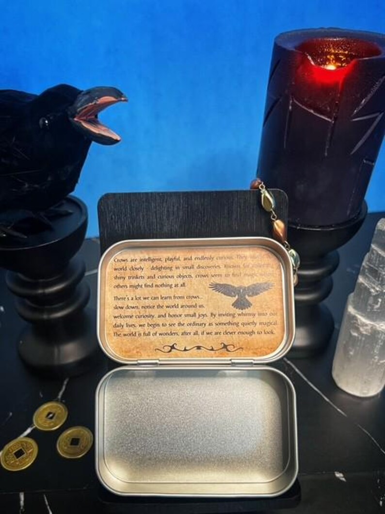 May include: An open, silver-colored metal tin with a printed message about crows, a black raven statue, a black candle, and three gold coins. The text on the tin reads, "Crows are intelligent, playful, and endlessly curious."