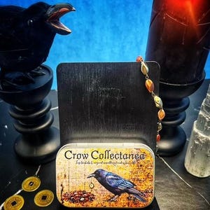 May include: A decorative tin with the text "Crow Collectanea" and a crow illustration. The tin is on a black stand, with a black crow statue, a lit black candle, and gold coins in the background. A strand of beads hangs from the stand.
