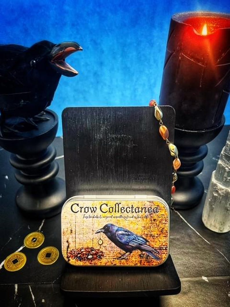 May include: A decorative tin with the text "Crow Collectanea" and a crow illustration. The tin is on a black stand, with a black crow statue, a lit black candle, and gold coins in the background. A strand of beads hangs from the stand.