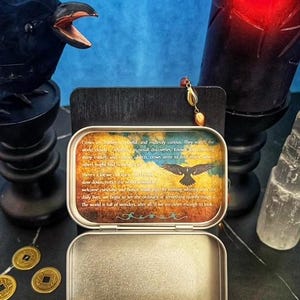 May include: An open, rectangular metal tin with a printed lid featuring a crow illustration and text. The tin sits on a black surface, with a crow statue, a candle, and coins in the background. A gemstone pendant hangs nearby.
