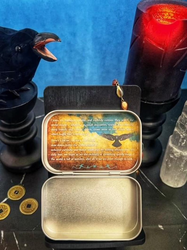 May include: An open, rectangular metal tin with a printed lid featuring a crow illustration and text. The tin sits on a black surface, with a crow statue, a candle, and coins in the background. A gemstone pendant hangs nearby.
