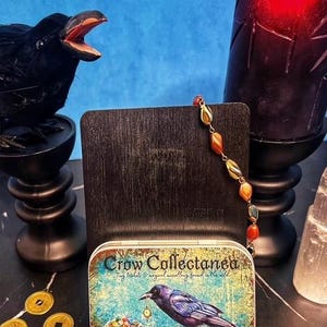 May include: A decorative tin with the text "Crow Collectanea" and a painting of a crow. A beaded necklace with orange and yellow stones is displayed next to the tin. A black raven statue and a red candle are also in the image.