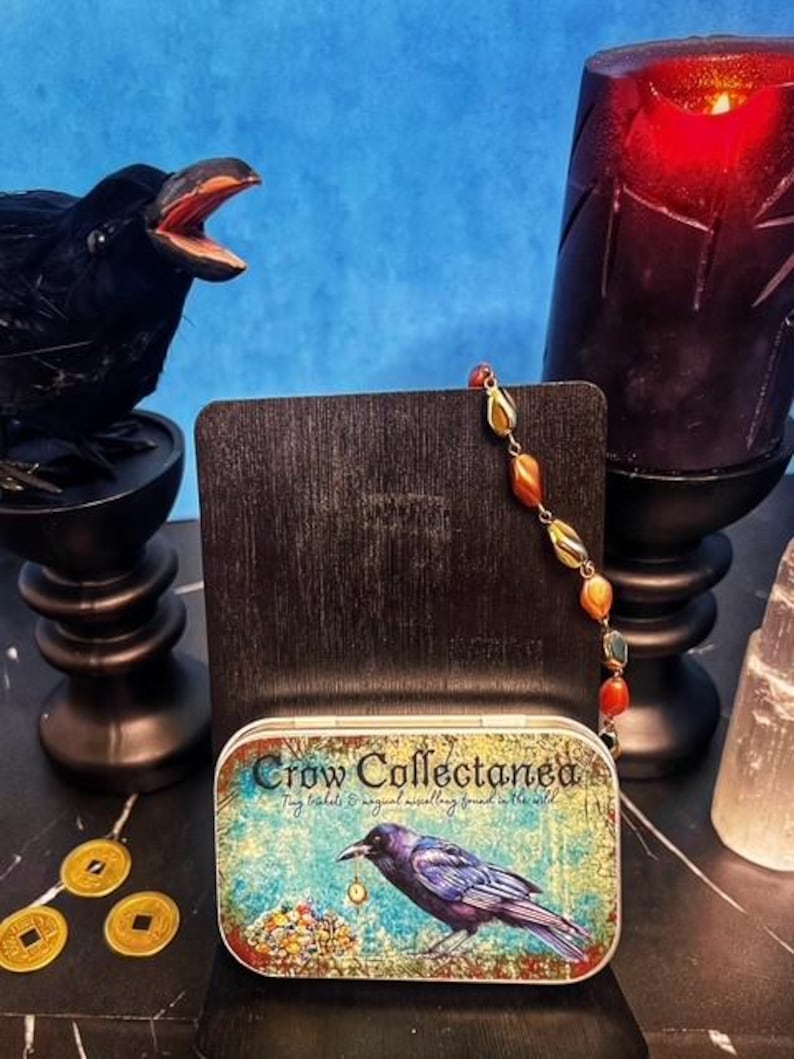 May include: A decorative tin with the text "Crow Collectanea" and a painting of a crow. A beaded necklace with orange and yellow stones is displayed next to the tin. A black raven statue and a red candle are also in the image.