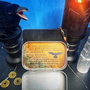 May include: An open metal tin with a printed message about crows, a raven statue, a lit candle, and gold coins. The tin's message includes text and a crow illustration. A clear crystal and a beaded strand are also visible.