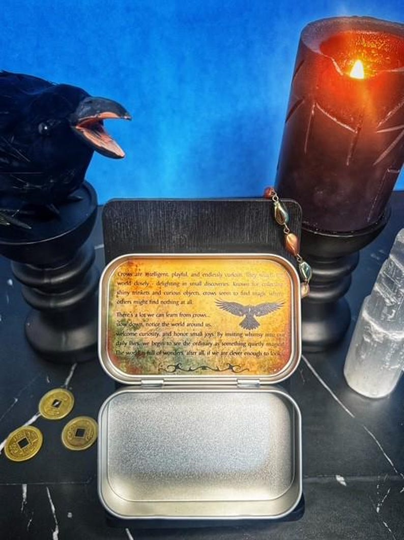 May include: An open metal tin with a printed message about crows, a raven statue, a lit candle, and gold coins. The tin's message includes text and a crow illustration. A clear crystal and a beaded strand are also visible.