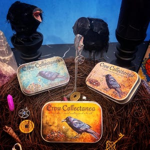 May include: An assortment of items with a gothic theme. Three rectangular tins with the text "Crow Collectanea" and a crow illustration are surrounded by decorative elements, including a black candle, crow figurines, and various charms.