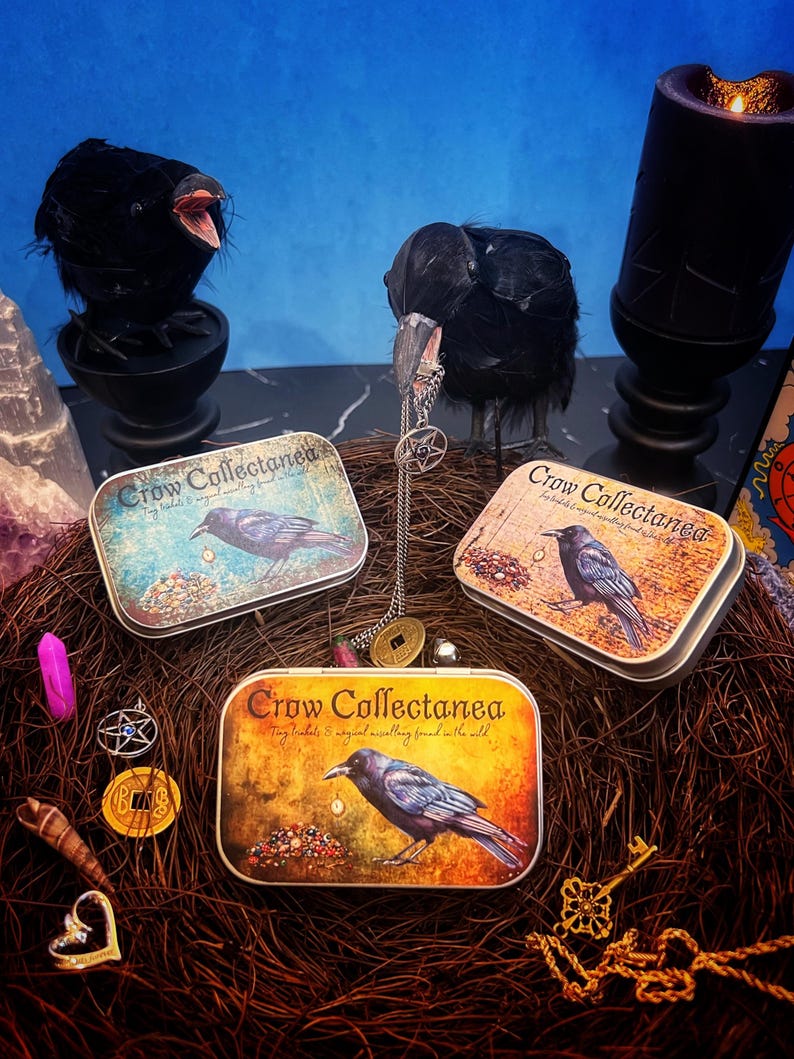 May include: An assortment of items with a gothic theme. Three rectangular tins with the text "Crow Collectanea" and a crow illustration are surrounded by decorative elements, including a black candle, crow figurines, and various charms.