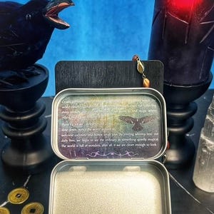 May include: An open, silver-colored metal tin with a printed message and a crow illustration. A black crow statue and a lit candle are in the background. Gold coins are in the foreground.