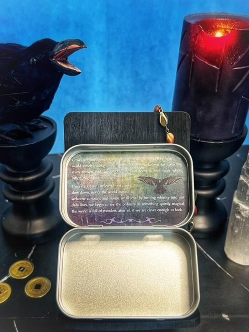 May include: An open, silver-colored metal tin with a printed message and a crow illustration. A black crow statue and a lit candle are in the background. Gold coins are in the foreground.