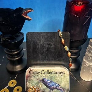 May include: A black crow figurine with an open beak sits on a black pedestal. A tin box with the text "Crow Collectanea" and a crow illustration is in front of a dark wooden stand. A beaded necklace and a red candle are also visible.